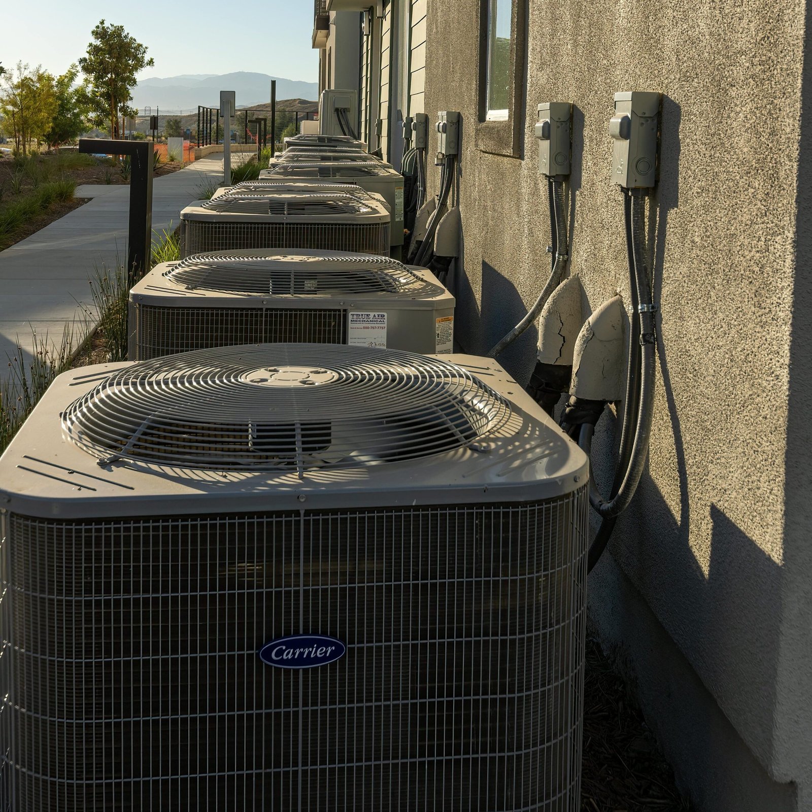 Line of air conditioning units on a city apartment exterior with a sidewalk view.